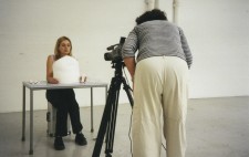 Mel sits behind a table, a model of the white corset in front of her, a bandage in her hand. Facing the table, Carol looks through the viewfinder of a tripd-mounted camera ready for a practice shoot.