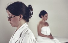 In the foreground Laura stands facing left of frame in a white coat. In the background, against a white backdrop, Isolte, wrapped in a white sheet, faces away from her.