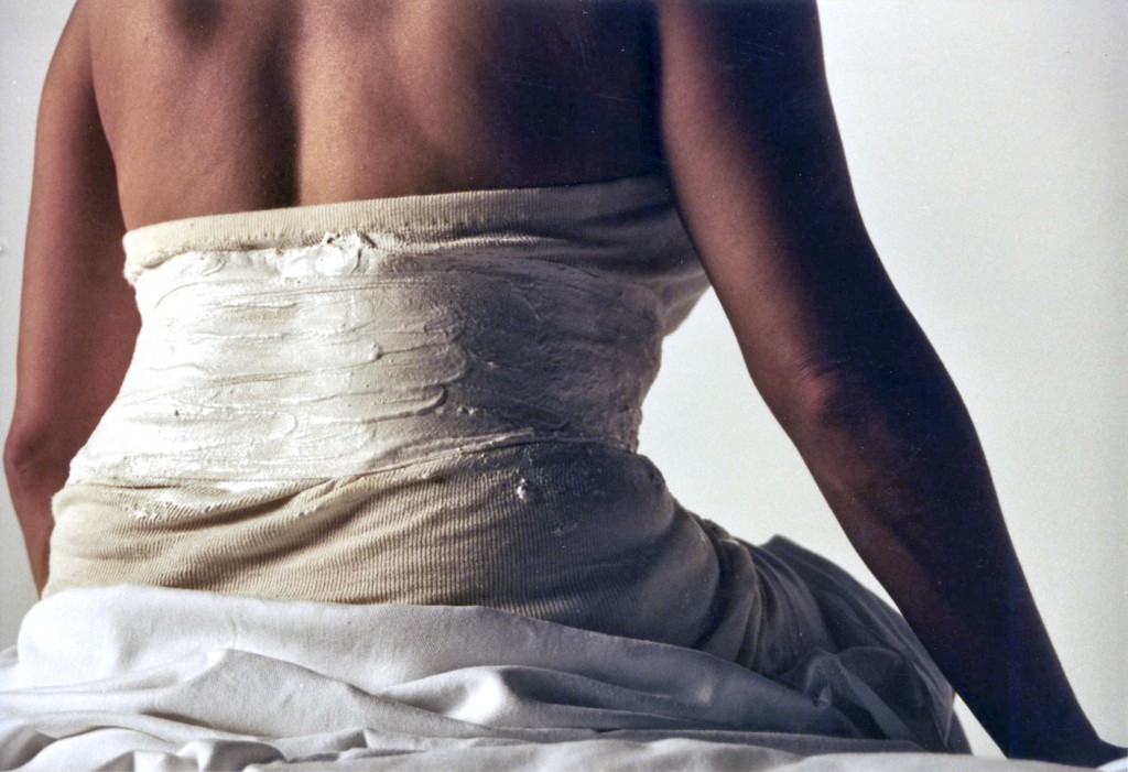 In close up, Isolte sits on a medical counch, her back to camera, her lower half draped in a white sheet and the unpainted corset constructed around her.