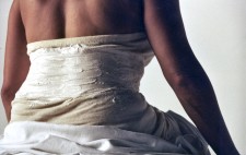 In close up, Isolte sits on a medical counch, her back to camera, her lower half draped in a white sheet and the unpainted corset constructed around her.