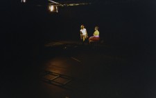 In a darkened studio, a pool of light illuminates Isolte who sits on a table in the distance. She wears a bright yellow top and purple skirt, talking to a crew member as scene is prepared.