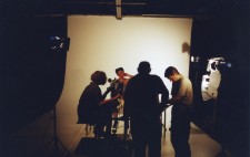 A studio filled with lights on stands. Against a stark white backdrop, Isolte faces forward, wrapped in the plaster corset. To the left of her sits Carol, who trains the camera on Isolte.