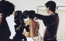 Carol adjusts her camera tripod, while a focus puller measures the distance from the camera to actor. Isolte sits facing a stark white backdrop, her back to camera and torso wrapped in a sheet, ready for the corset to be applied.