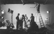 A monochrome photograph: a studio is filled with lights on stands, ladders and boxes of film kit. Against a white backdrop, Frida (Isolte Avila) faced forward, wrapped in the white plaster corset.  The camera is trained on Isolte, the crew in place. On a tall ladder, Mel holds a small pot extended on a pole high above Isolte's head.