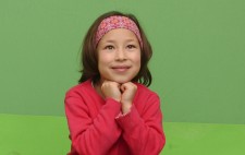 Maia sits on a low stool against a uniformly bright green screen and floor. She wears a red top and skirt, a flowery headband and purple tights and battered old trainers. Her elbows rest on her lap and her chin on her hand. She gazes dreamily upwards, a small smile on her face.