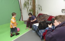 A boy of around five, wearing a bright orange flowery shirt and jeans poses against a greenscreen ready to kick a ball. Liz faces him, demonstrating how she wants him to place his arms.