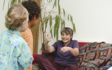 Two boys of around four years old are dressed in brightly coloured clothes and stand poised on the greenscreen floor. On the sofa, facing them, is Liz who uses her hands to demonstrate the movement she wants from them.