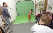 Director Liz Crow gives directions to a boy dressed in a multicolored rainbow t-shirt and brown trousers who stands in front of a greenscreen. He carries a long fabric ribbon in rainbow colours which is being held aloft at the other end by Bryan to look as though it is streaming in the breeze. Matt has his back to the camera.