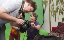 Matt and Liz look intently at the display on Matt’s camera. Liz points at the image whilst one of the child actors can be glimpsed in the background peeping in.