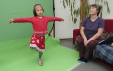 Maia stands on one leg with her arms outstretched, looking dreamily, in front of a greenscreen. Liz sits on a sofa just to the side.