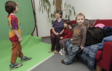 Against a greenscreen, a boy of about six stands in position, looking towards Liz who is talking to him. Two of the other children sit next to her on the sofa.