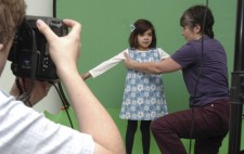 Against a greenscreen, a girl of around five years old stands facing towards the camera dressed in a white top and blue flowery pinafore, a blue ribbon in her hair. Liz kneels to her side, gently moving her arms into position.