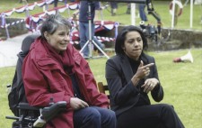 At the lakeside, Director Liz Crow sits in her wheelchair in a red raincoat and scarf, a sign language interpreter seated next to her. In the background, the jimmy jib is being prepared for the filming the big race.