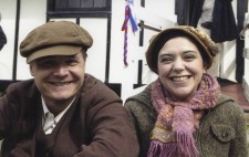 At the lakeside, James and Poppy beam at the camera. James is dressed in a brown suit and cap, whilst Poppy wears a vintage hat with a modern sweater and scarf covering her 1930s summer dress.