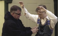 Jacob stands, arms raised, as Paul fits his microphone ready for the market scene. Dressed in shift and tank top, with his fruit-and-veg stall apron, Jacob is fitted with the vintage bakelite hearing aid.