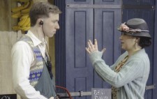 Next to a green and red wooden barrow, laden with fruit and vegetables, Walter and Gloria stand facing each other. She wears a 1930s floral dress and light blue cardigan, and a small hat with flowers. She holds her hands aloft, signing hesitantly. Walter wears a greengrocer’s apron and stands with his thumbs tucked in the front pocket, mesmerised by Gloria and her signing.