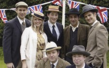 Seven members of the cast from the lakeside shoot pose together for a photo, all dressed in period costume, Union flag bunting festooned behind them. The men wear suits and the woman is wearing a yellow dress and a white overcoat. All wear boaters, fedoras or caps.