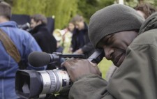 Cameraman David Ellington looks through the camera viewfinder, members of the crew busy in the background.