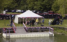 A white marquee stands behind the swimming pontoon, which is decorated but still empty of actors. The crew are on the banks preparing to shoot, with camera tests taking place.