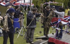 Members of the crew stand in a line by the pontoon holding feeding out lane ropes ready to divide the lanes in the swimming lake. Behind them, the scene is already decorated in festive red, white and blue bunting.