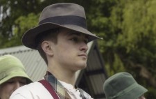 A cast member waits on set between lake scene takes, a loose tie around his neck and a brown fedora on his head.