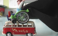 A Playmobil woman sits on a wheelchair perched on top of a miniature red model VW van, with ‘love and lib’ painted onto its side. The woman’s arms are flung high and, from her shoulders, a piece of black fabric flows behind her.
