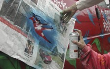 A close up of a hand holding a newspaper stencil of a silhouetted bird against the side of a red VW camper van. Another hand sprays aerosol paint onto the paintwork to add a bluebird to the leaves and sprays of white flowers already adorning the vehicle.