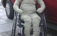 A life-sized hessian fabric doll with no features sits naked on a manual wheelchair next to a red van.