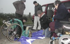 In early morning light in open country, 'Katie', the life-sized fabric doll is clothed in long green dress and flowing blonde wig, a flower in her hair, is seated on a wheelchair tipped backwards to the ground. Members of the crew, dressed against the cold, attach a swathe of blue fabric to her shoulders.