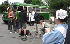 Photograph: A green vintage bus is in the background, a camera set up and crew busily preparing for a shot. Two white-coated orderlies stand near the bus. Terry looks skywards. In the foreground the nurse holds a cup of tea and watches the preparations.