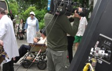 Photograph: In foreground, Alex balances a heavy camera on one shoulder. Behind him the inmate actors assemble ready to be herded from institution doors and into the bus. One man sits on a 1930s canvas-seated wheelchair.