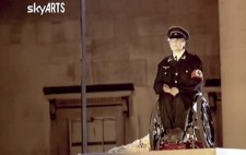 A close-up screenshot, the SkyARTS logo in the top left corner, shows Liz on her wheelchair on the plinth and wearing a black Nazi uniform.