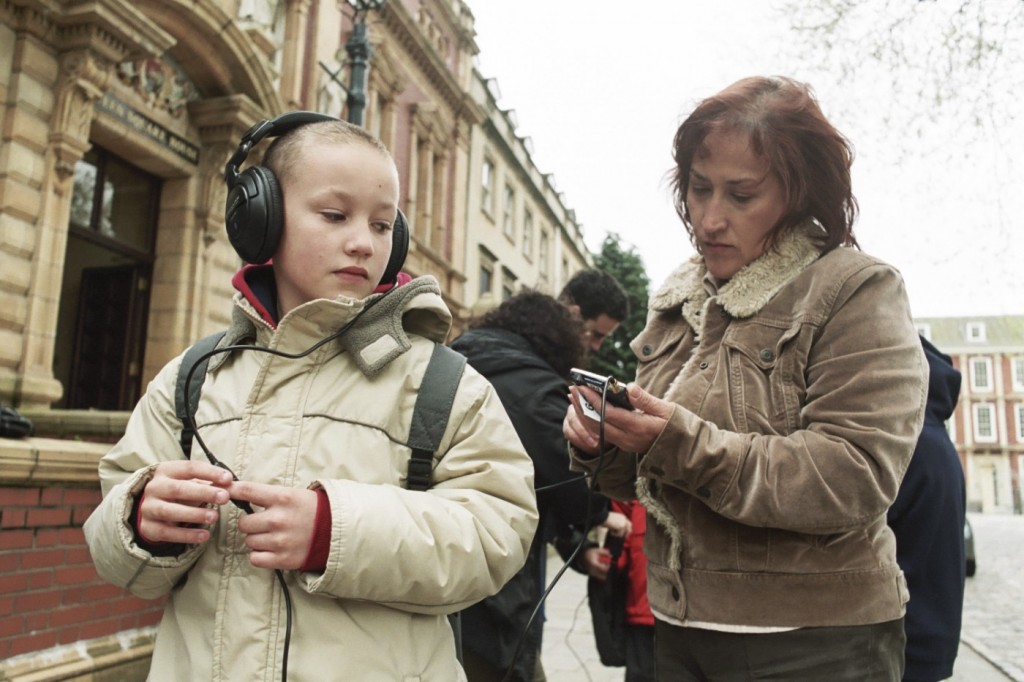 A primary age boy stands at the edge of the Square wearing a backpack and large earphones, whilst Jo sets the handheld computer.