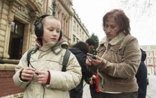 A primary age boy stands at the edge of the Square wearing a backpack and large earphones, whilst Jo sets the handheld computer.