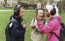 Three primary age children stand in the grassy Square, wearing large earphones and excitedly exchanging their experiences of 1831 Riot!