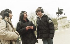 Three primary age girls stand on gravel path in the Square, wearing large earphones and sharing their experiences of Riot. In the background is a plinth with a statue of a figure on horseback.