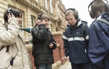Four primary age school children wearing large earphones stand together at the edge of the Square listening to the start of 1831 Riot!