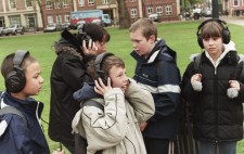 Four primary age children and a teacher stand in a grassy area of the Square wearing large earphones, each absorbed in the audio and looking to different areas of the Square. 