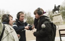 Three primary age school children wearing large headphones gather in Queen's Square near a statue of a figure on a horse, animatedly compare experiences as they listen to the audio.