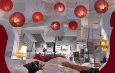 A montage of photos giving a panoramic view of Liz's view from the bed: two standard lamps frame the bed with a warm yellow glow, the space extends beyond the foot of the bed to a cafe area, the words 'cafe' lettered on a large picture window. Above the bed is a fabric canopy in white, festooned with multiple red paper lamp shades. Liz's arm extends across the sheets.