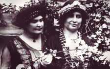 A portrait of the two women in mid-life. They are elegantly dressed in late Edwardian fashion, both wearing hats, and standing against a backdrop of a hedge of white blossom.