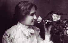 Helen wears a white Edwardian blouse with wide sleeves and a tie bow at her neck. With her left hand, she pulls a rose to smell, while her right rests upon a Braille book on her lap.