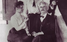 Helen and Mark Twain (Samuel Clemens) sit on chairs. He faces the camera and she is turned towards him. Helen wears a floor-skimming dark skirt and a white blouse with high collar. Behind her stands Annie, her hand resting on Helen's raised palm to fingerspell the conversation. Behind Twain, another (unidentified) man stands, pipe in hand. The photograph is signed 'SL Clemens.'