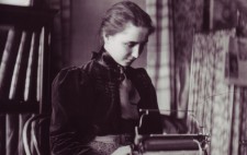 Helen sits at a desk before a Braille typewriter. Her hair is pulled back from her face into a braid and she wears a dark blouse with a high next and large bow.