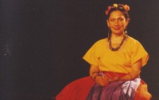 Against a pitch black backdrop and seated on a black bench, Isolte is dressed in a bright yellow, short-sleeved shirt and long, flowing, orange skirt, with a purple sash. Brightly coloured flowers are woven into her hair and she is adorned with jewellery: large, chunky earrings, necklace, bracelets and rings.