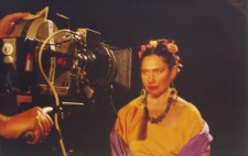 A close up of Isolte, dressed in bright yellow top, with a purple shawl around her shoulders and flowers in her hair. A large film camera is focused on her as she sits against a pitch black backdrop and looks directly into the lens.