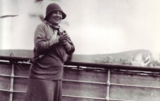 Standing at a ship's rail, land in the background and a seagull overhead, Helen smiles broadly and holds her hands together in front of her chest. She is dressed in late 1930s fashion, a calf-length skirt, silk scarf tied around her neck, a cloche hat and sturdy shoes.