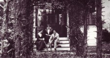 The exterior of a New England clapperboard house on three storeys, with a second storey balcony. Helen, Annie and John sit at ground level with their dog on steps leading onto a porch.