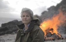 Liz Crow looks to camera. Behind her, the bonfire roars in orange flame against a backdrop of beach shingle and bolders.