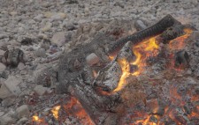 The embers glow, the last few flames flickering. At the out edges, the figures are visible, encased in chicken wire, merging into the surrounding shingle.